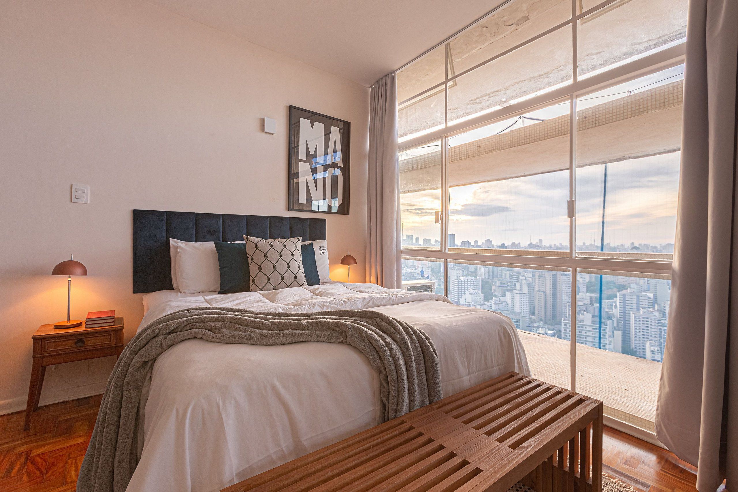 Une chambre avec une grande fenêtre, un lit, une table de chevet avec une lampe, et une vue sur le paysage urbain.