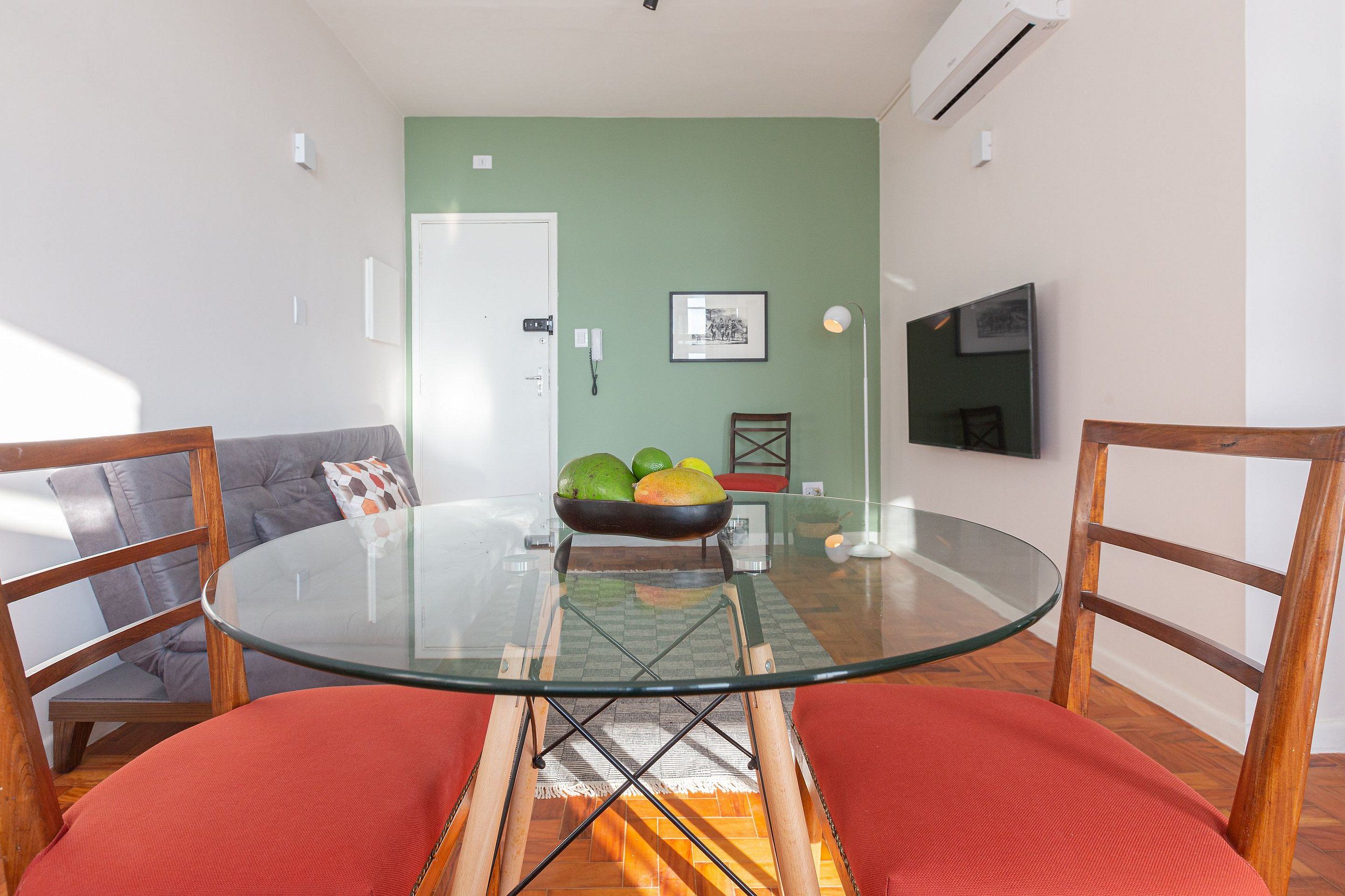 A dining area with a glass table, red chairs, a bowl of fruit, and a green wall.