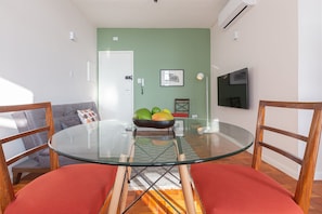 Un coin repas avec une table en verre, des chaises rouges, un saladier de fruits et un mur vert.