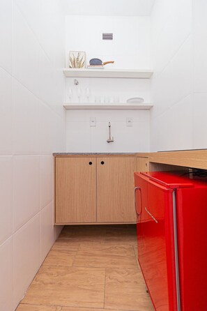 A compact kitchen with white tiles, a wooden cabinet, a red appliance, and a shelf with glasses and a decorative item. - Vossa Bossa Copan (São Paulo)