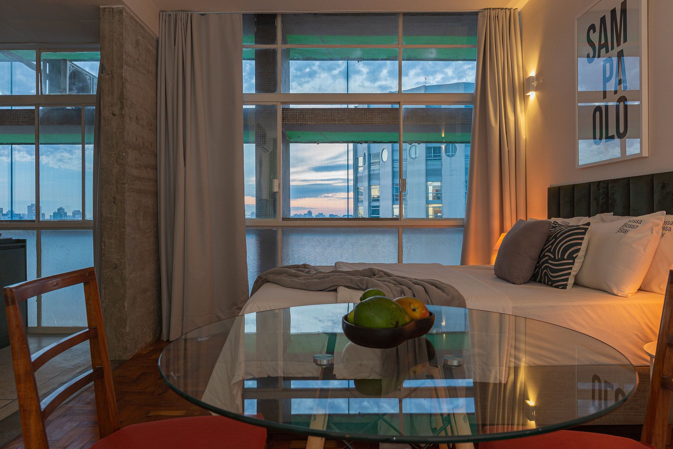 Een moderne hotelkamer met een glazen eettafel, een comfortabel bed met kussens en uitzicht op de stad door grote ramen.