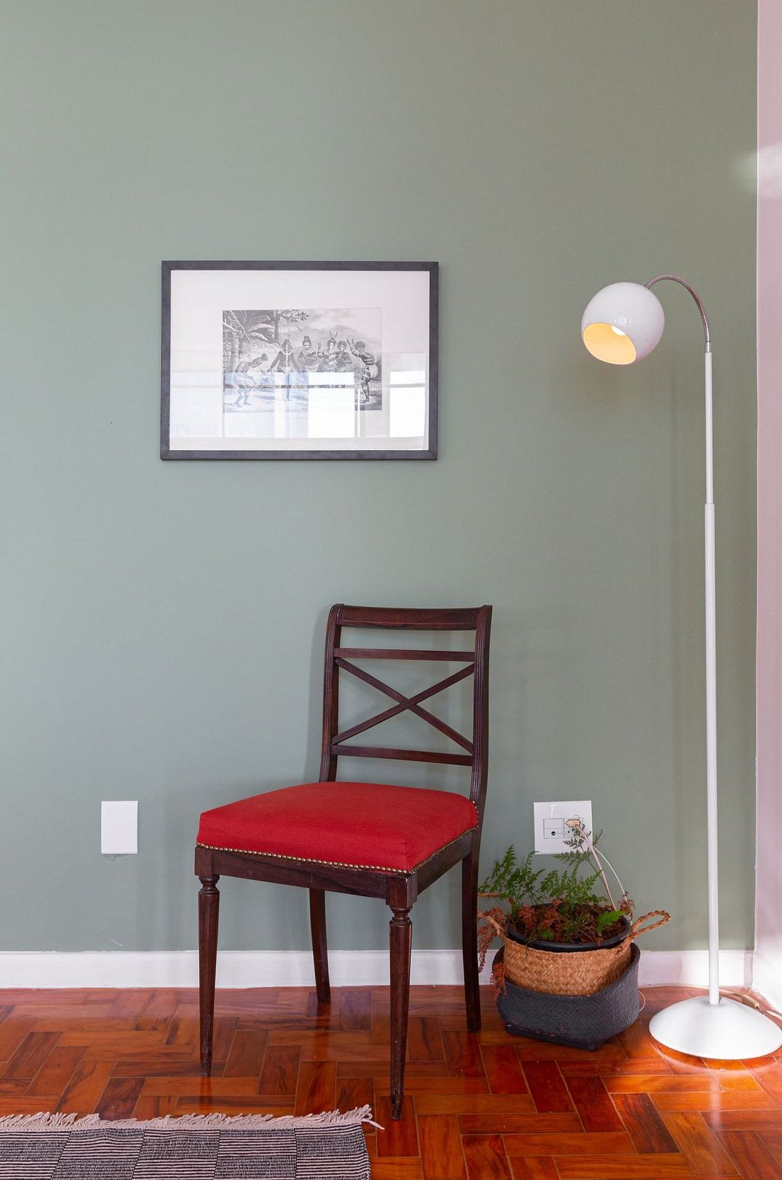 Ein Holzhocker mit rotem Kissen, ein gerahmtes Bild an der Wand, eine Stehlampe und ein Blumentopf auf dem Boden.