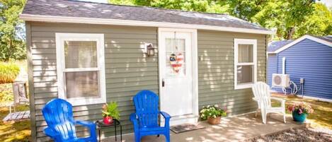 A small single-story house with two blue Adirondack chairs outside, a white door with a heart-shaped decoration, and a small porch area with potted plants.