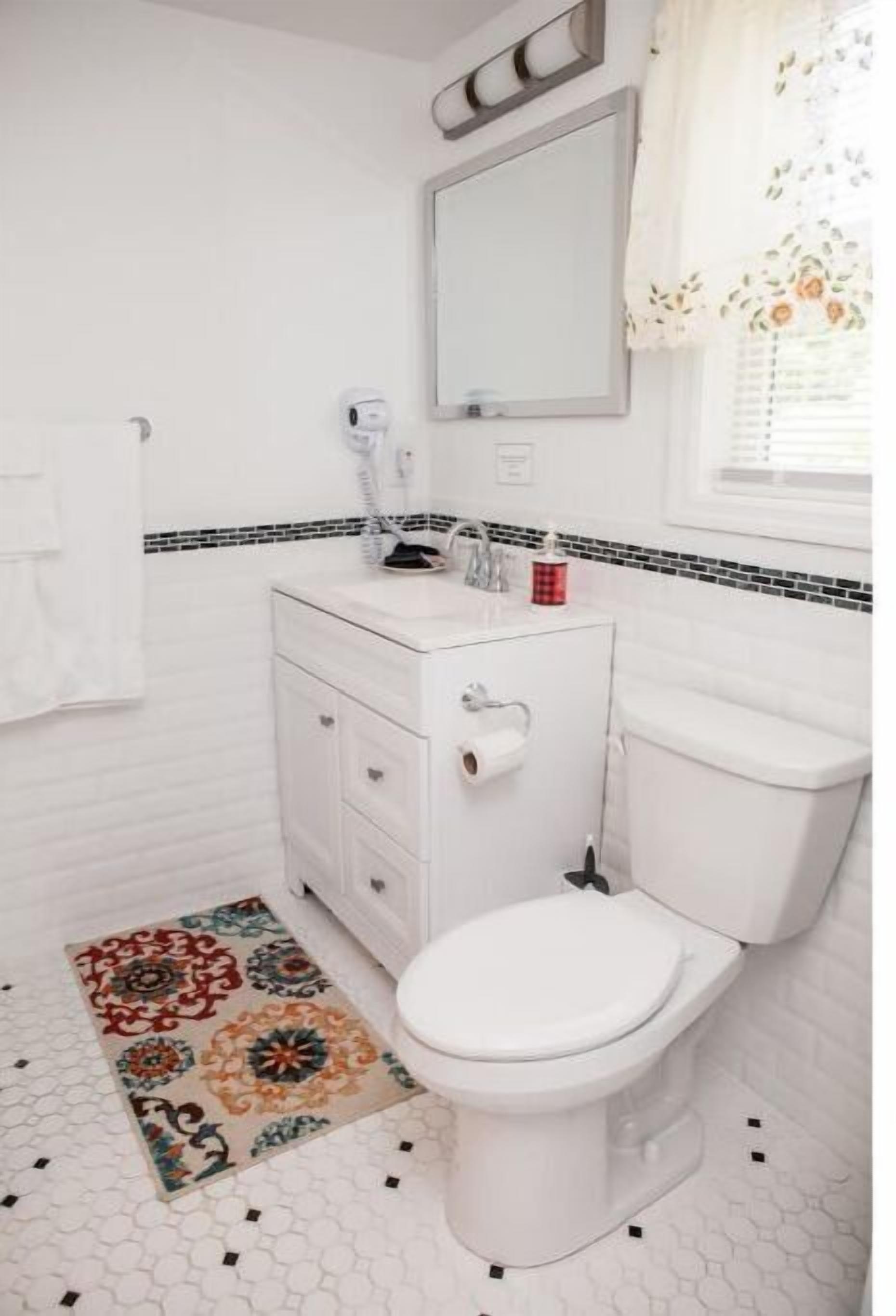 Un baño con inodoro blanco, un lavabo blanco con espejo arriba y un tapete estampado en el suelo.