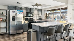 A modern kitchen with stainless steel appliances, a central island with bar stools, and a view through large windows. - PuttPutt, Pickleball, Basketball, Ping Pong & Game Room! Pool & Spa! (Scottsdale)