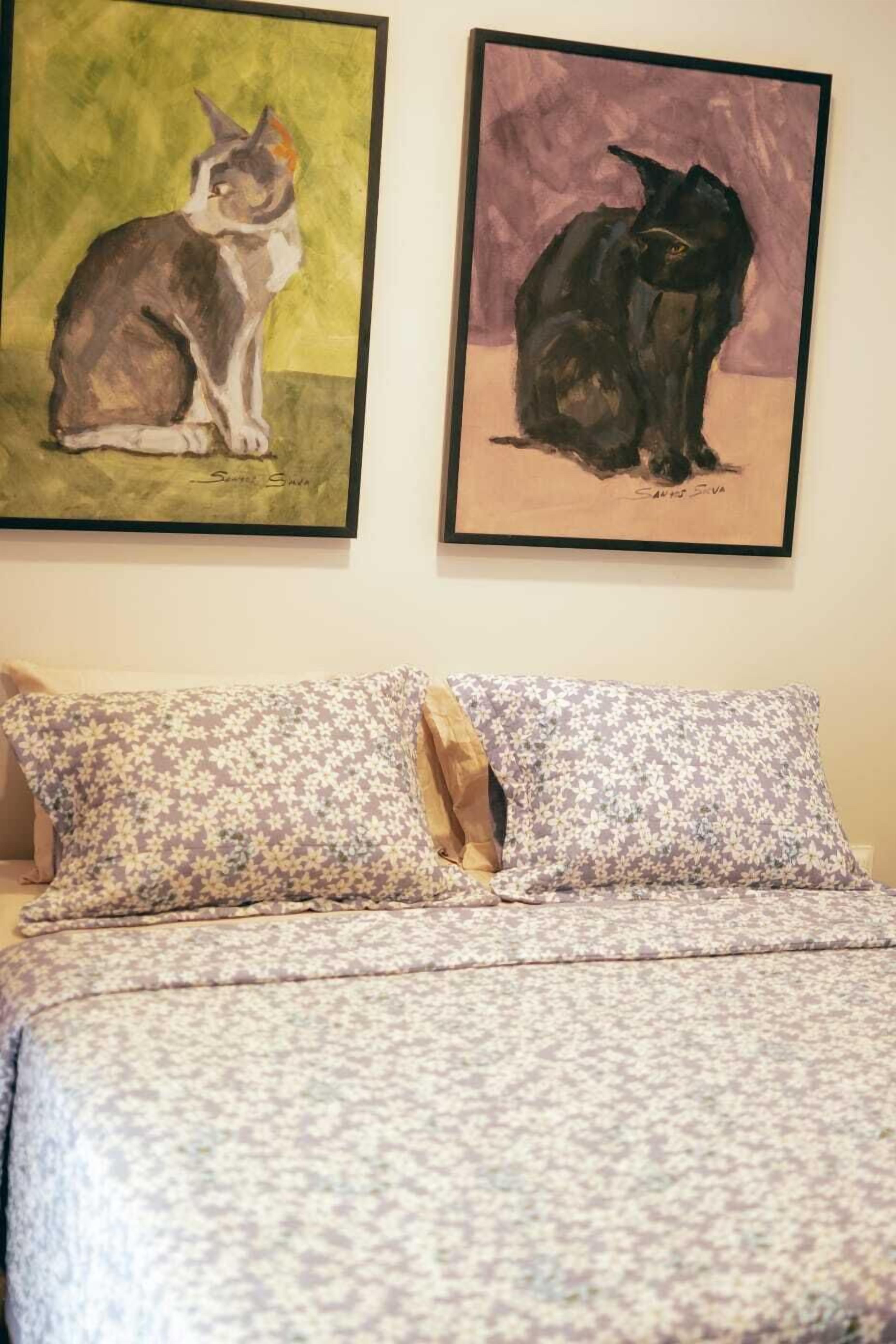 A bedroom with a bed, two framed paintings of cats, and floral-patterned pillows.