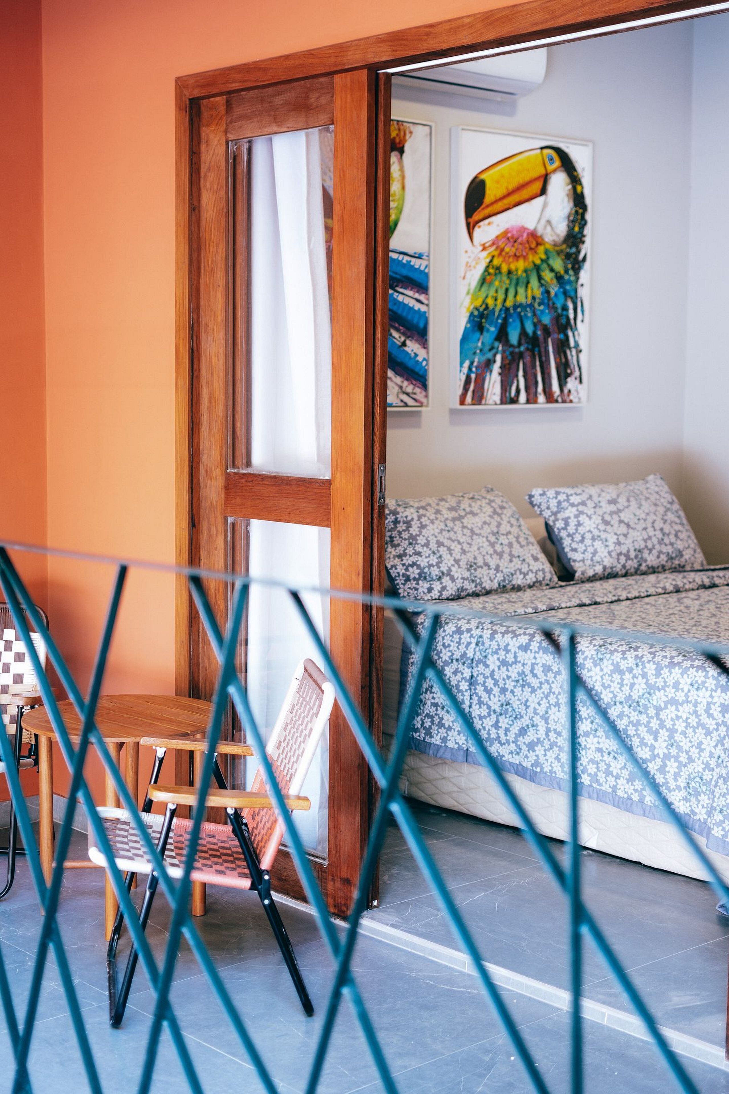 Ein Schlafzimmer mit einem Bett, einer Holztür, einem Fenster und einem farbenfrohen Porträt eines Tukans an der Wand.