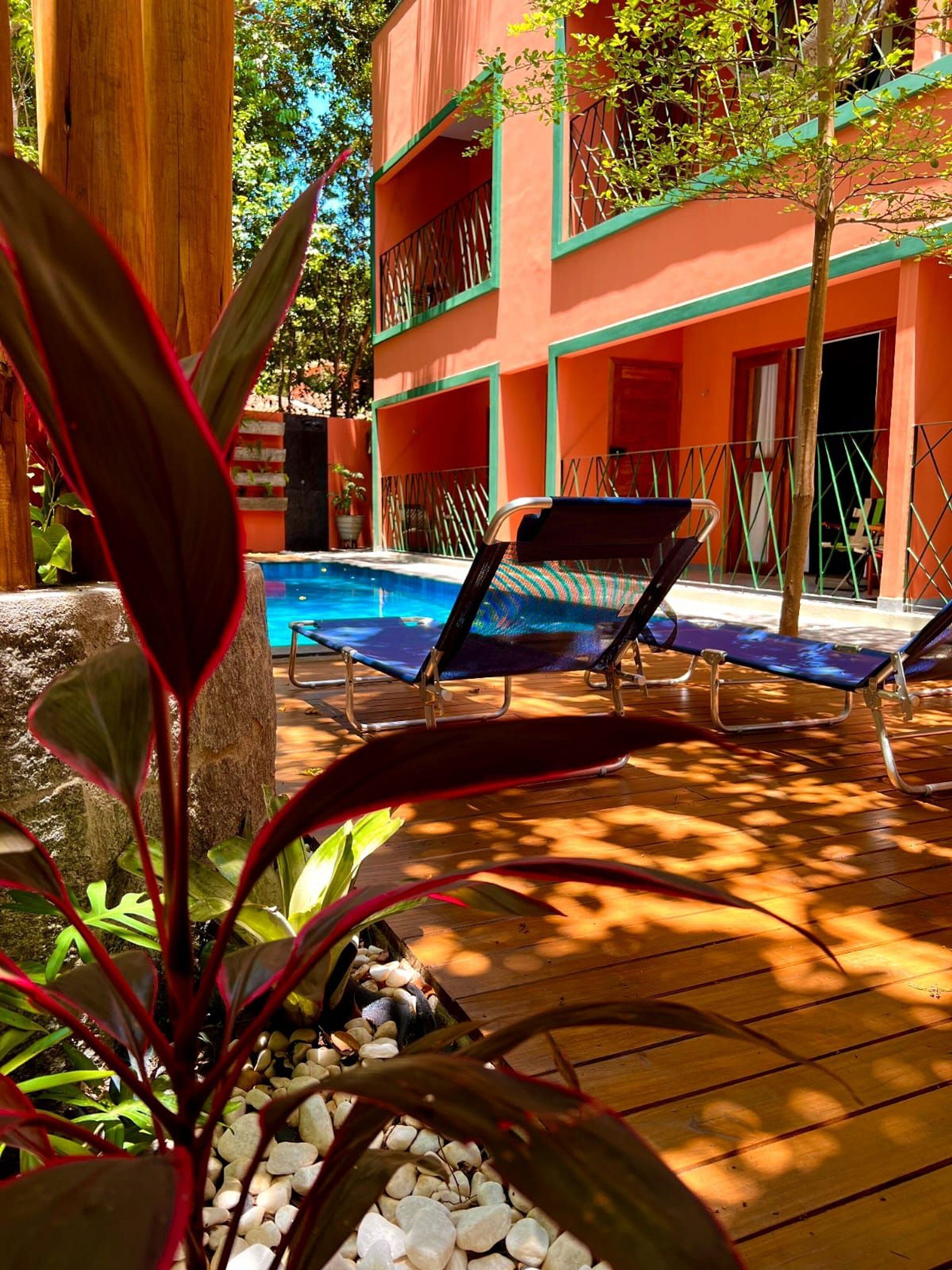 Un área junto a la piscina con sillones de descanso, un edificio con balcones y una planta tropical en primer plano.