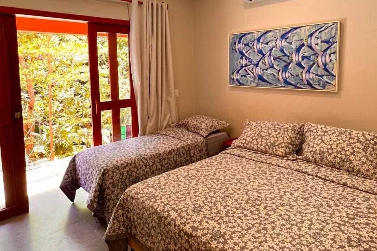 A bedroom with two beds, a window with curtains, an air conditioning unit, and a painting on the wall.