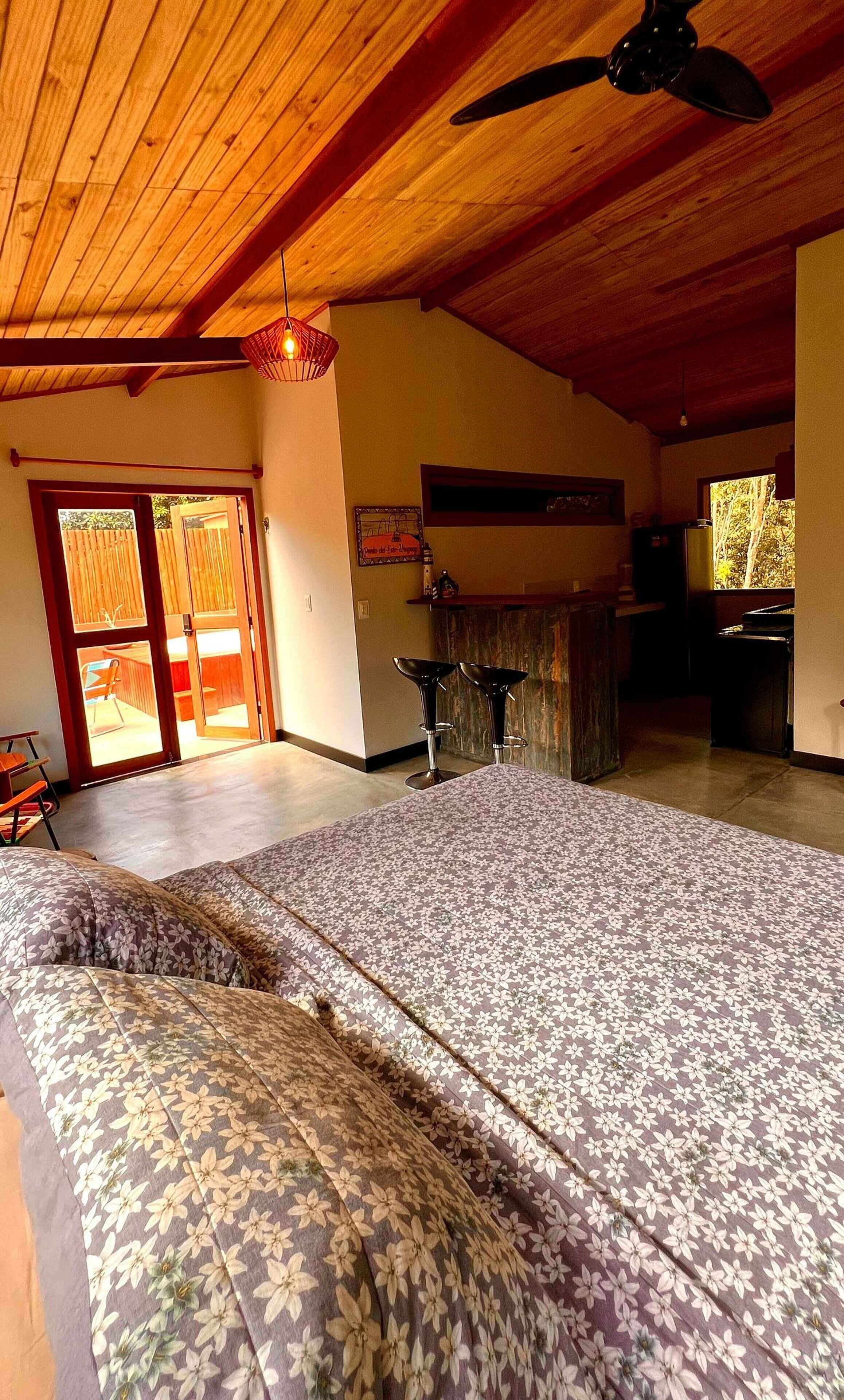Une pièce spacieuse avec un plafond haut, un ventilateur de plafond, un grand lit avec une literie à motifs floraux, un coin bar et une vue sur l’extérieur.