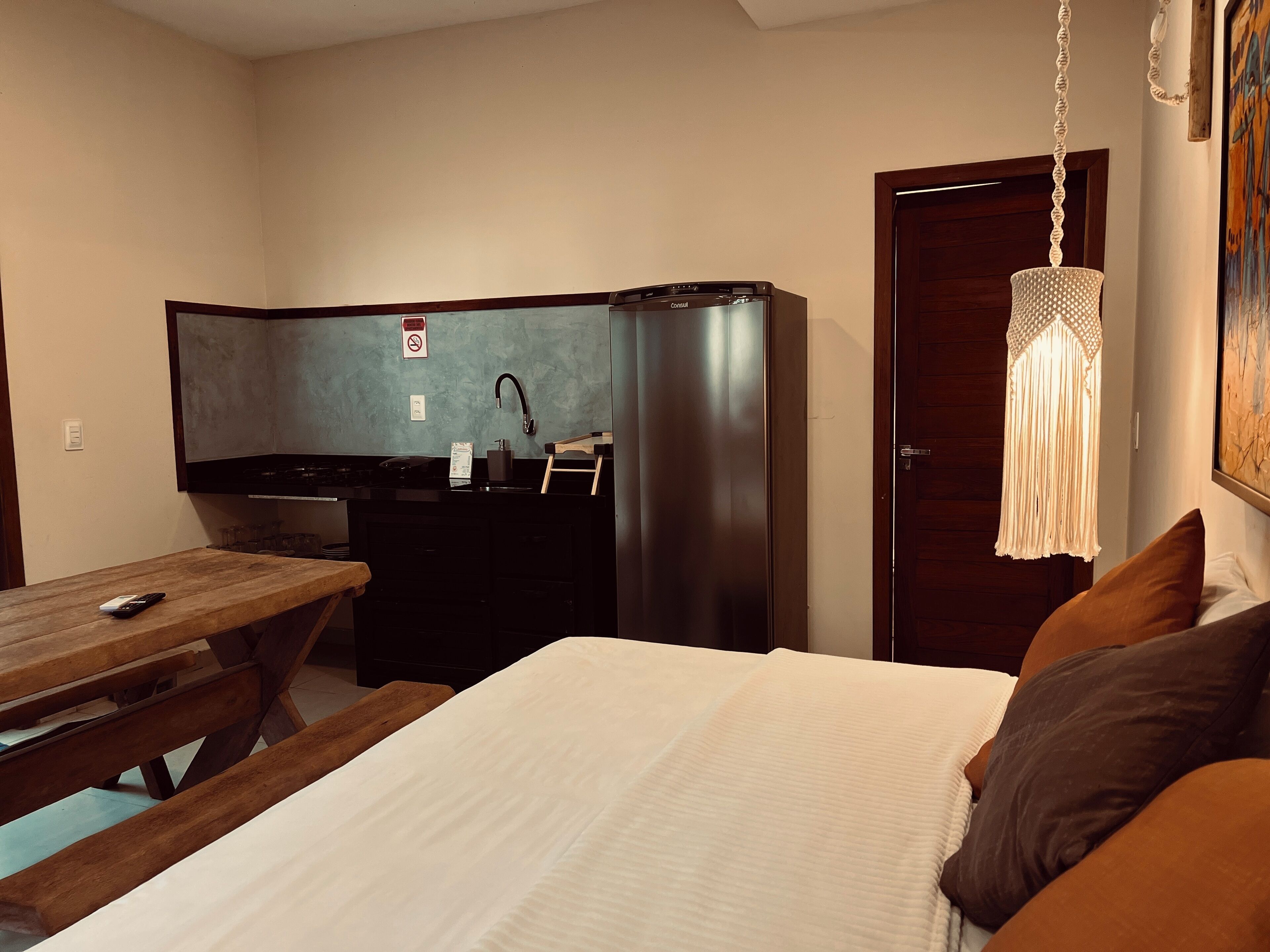 A compact hotel room with a bed, a wooden table, a kitchenette, and a hanging light fixture.