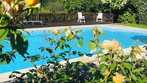 Uma piscina rodeada por vegetação exuberante e flores brancas, com duas espreguiçadeiras brancas e uma cerca de madeira ao fundo.