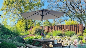 Uma mesa de piquenique de madeira com um guarda-sol branco, rodeada por um jardim com diversas plantas e flores.