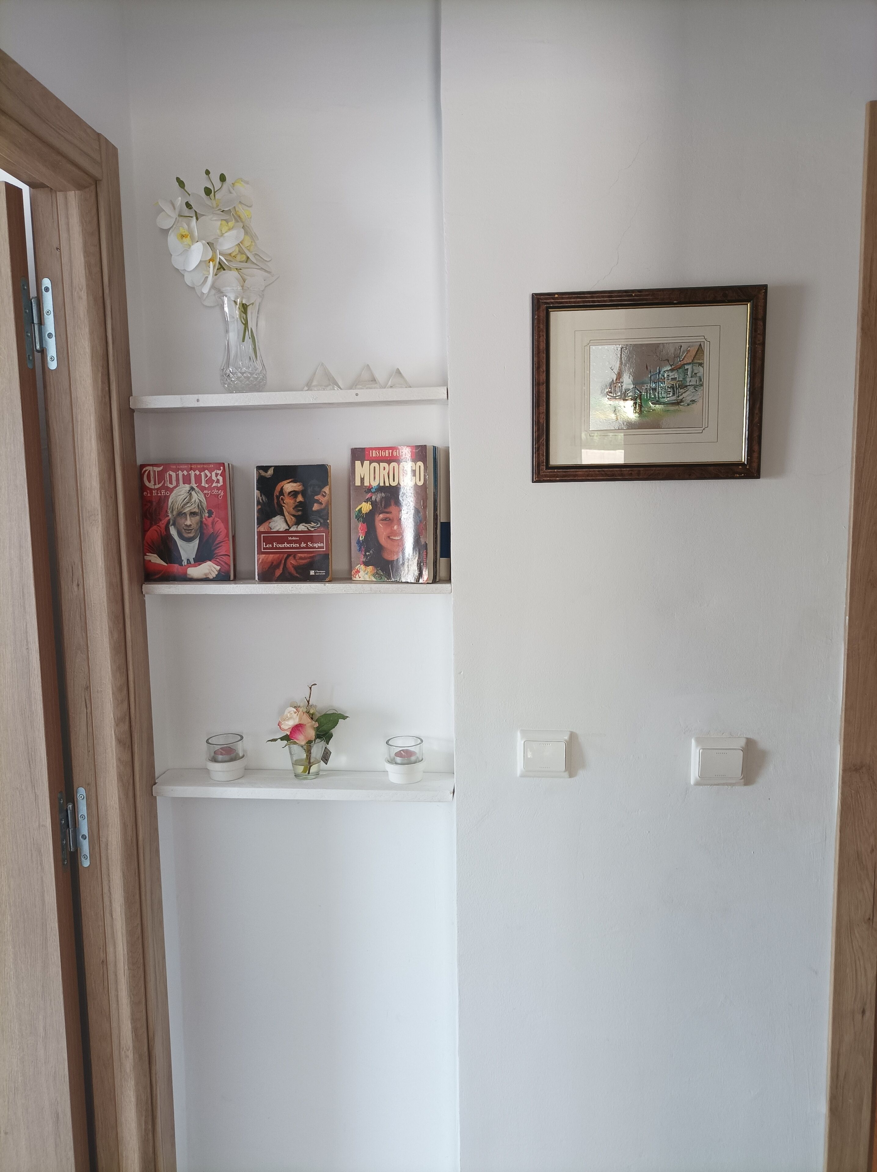 Ein Zimmer mit Holztür, einem weißen Regal mit Büchern und einem Blumenstrauß, einem gerahmten Bild an der Wand und einem Thermostat.