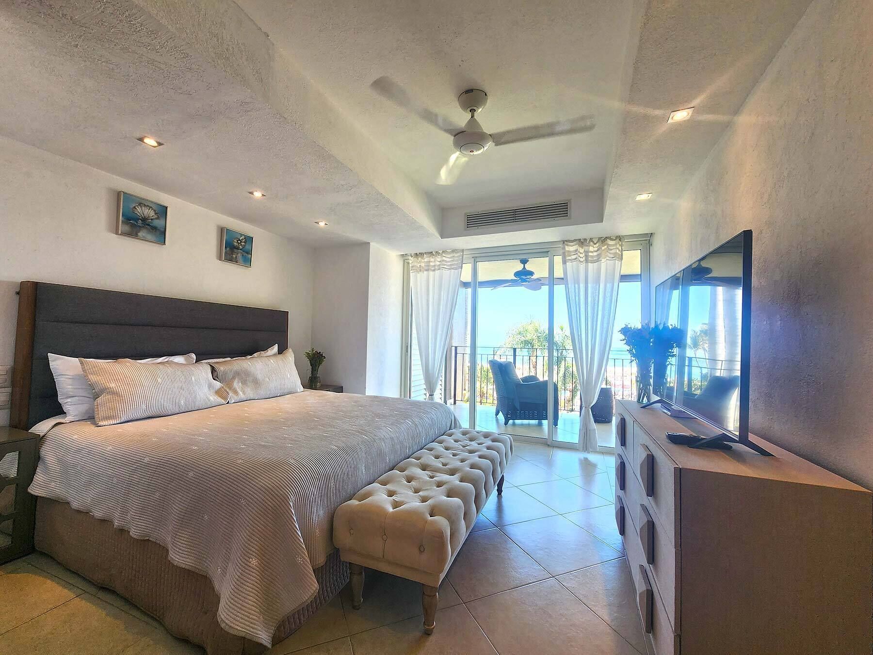 Un dormitorio con una cama grande, un banco, un ventilador de techo, un televisor y vistas al exterior.
