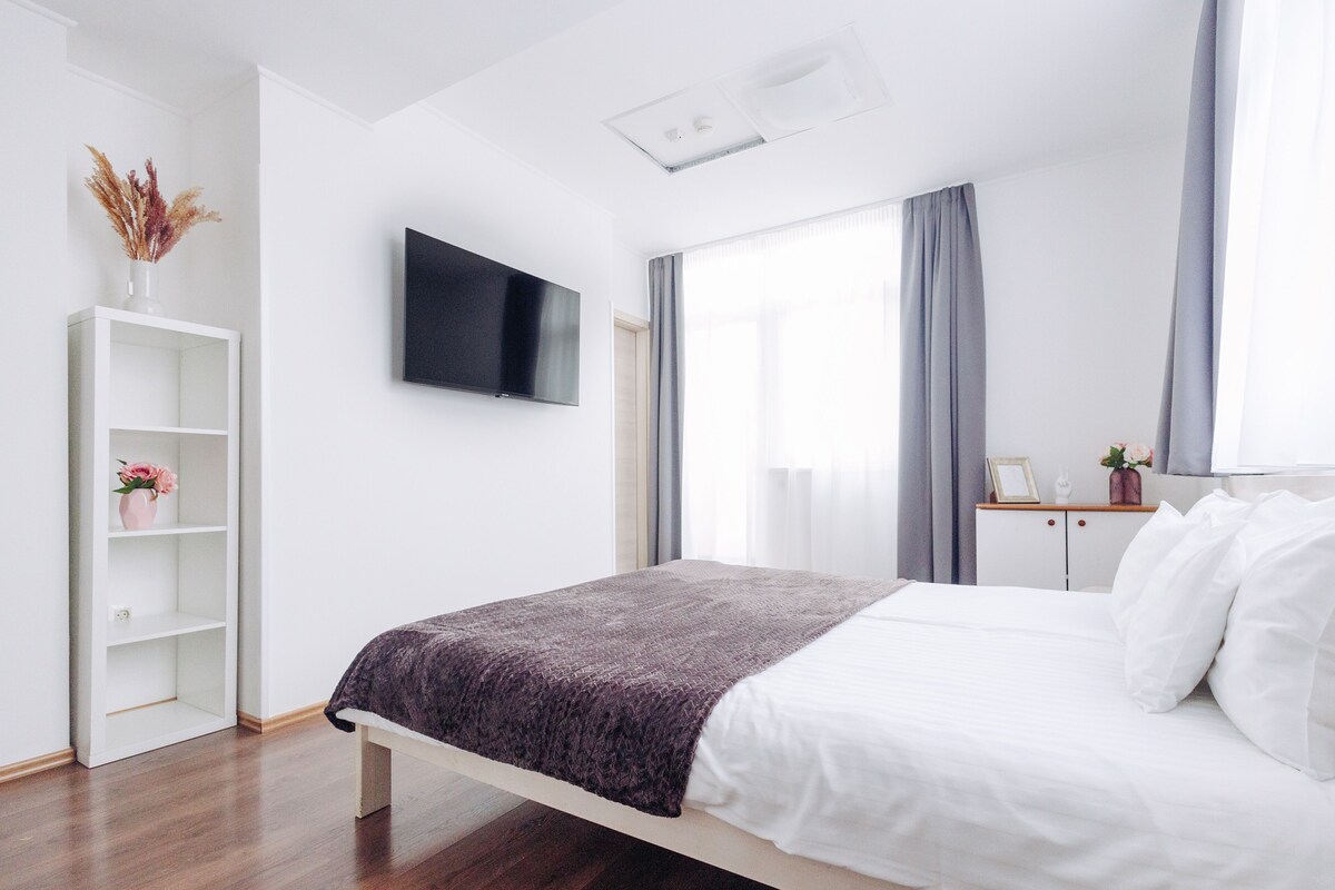 Une chambre moderne avec un lit, un téléviseur fixé au mur, une étagère blanche et une fenêtre avec des rideaux.