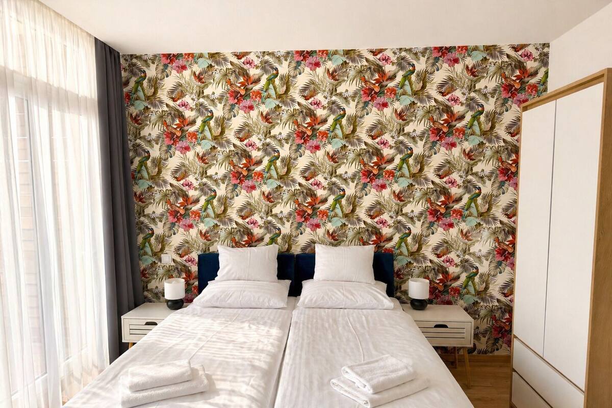 A double bed with white linens and towels, a nightstand with a lamp, and a tropical floral wallpaper.