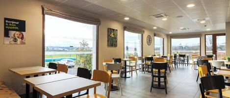 A spacious cafe with multiple tables and chairs, large windows offering a view of the outside, and a ceiling with a unique pattern.