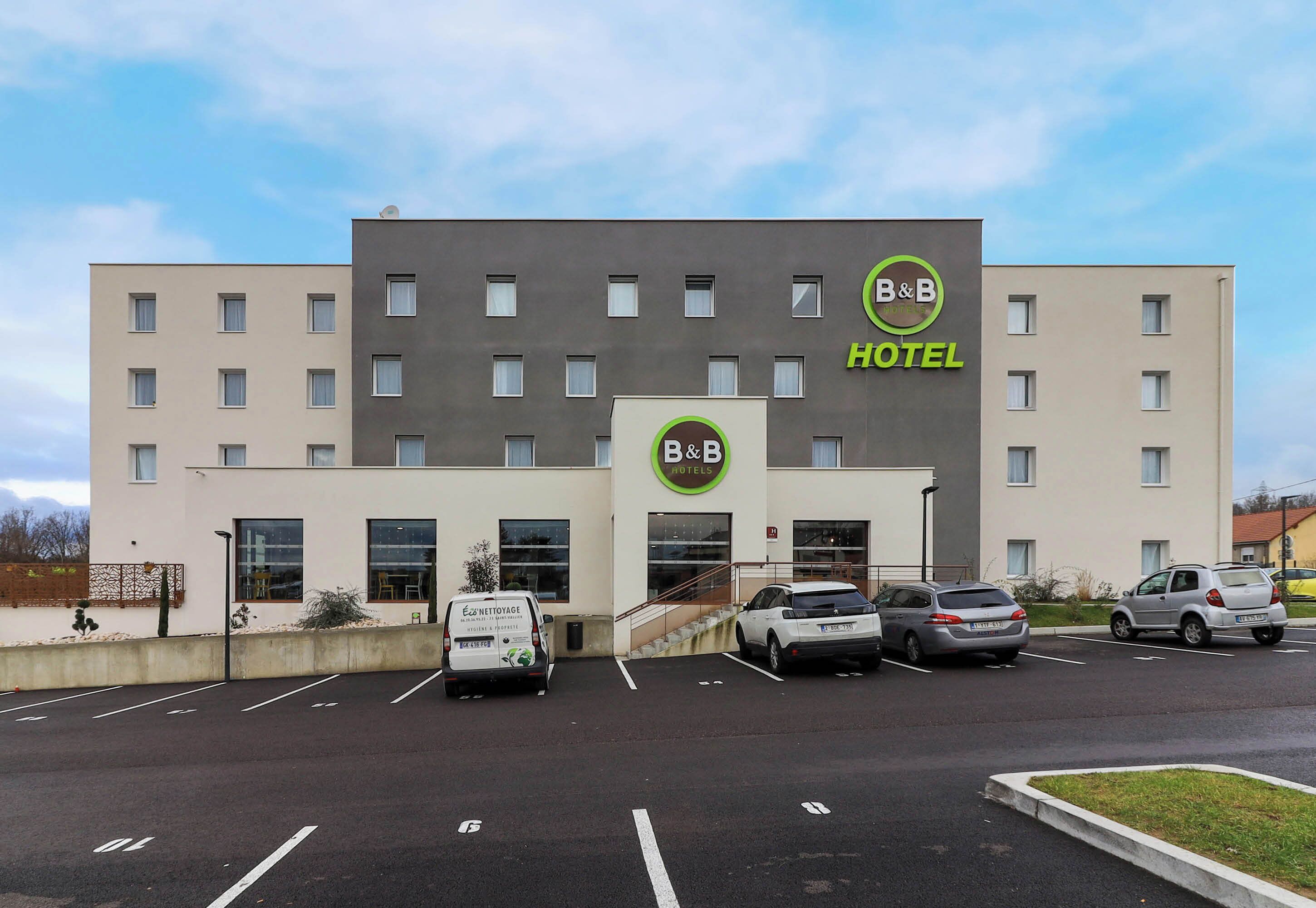 Un hôtel-restaurant de type « bed and breakfast » doté d’une façade moderne, d’un parking où sont garées plusieurs voitures et qui bénéficie d’un ciel dégagé.