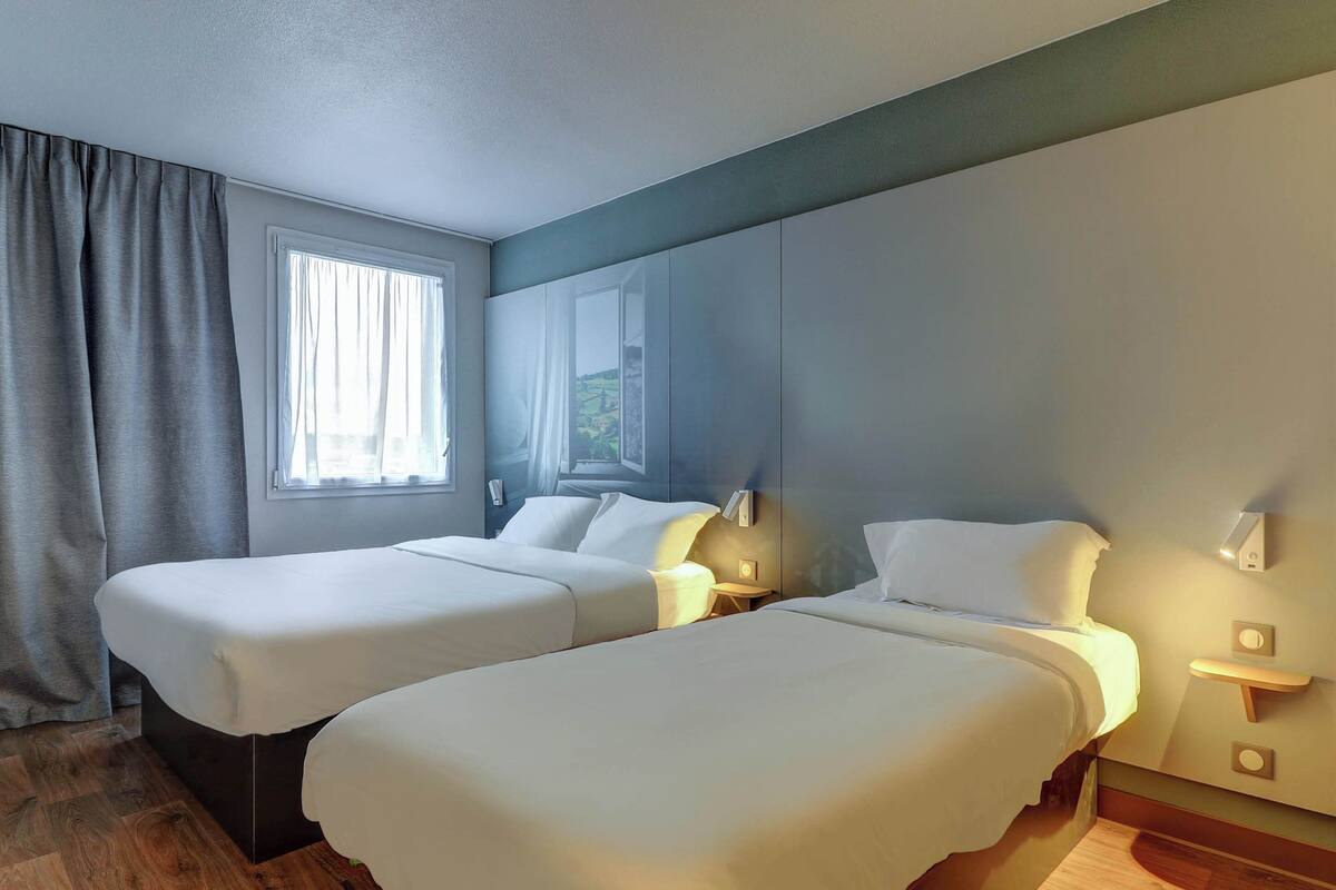 Ein Hotelzimmer mit zwei Betten, einem Fenster mit Vorhängen, einem Nachttisch mit Lampe und einer Wandleuchte.