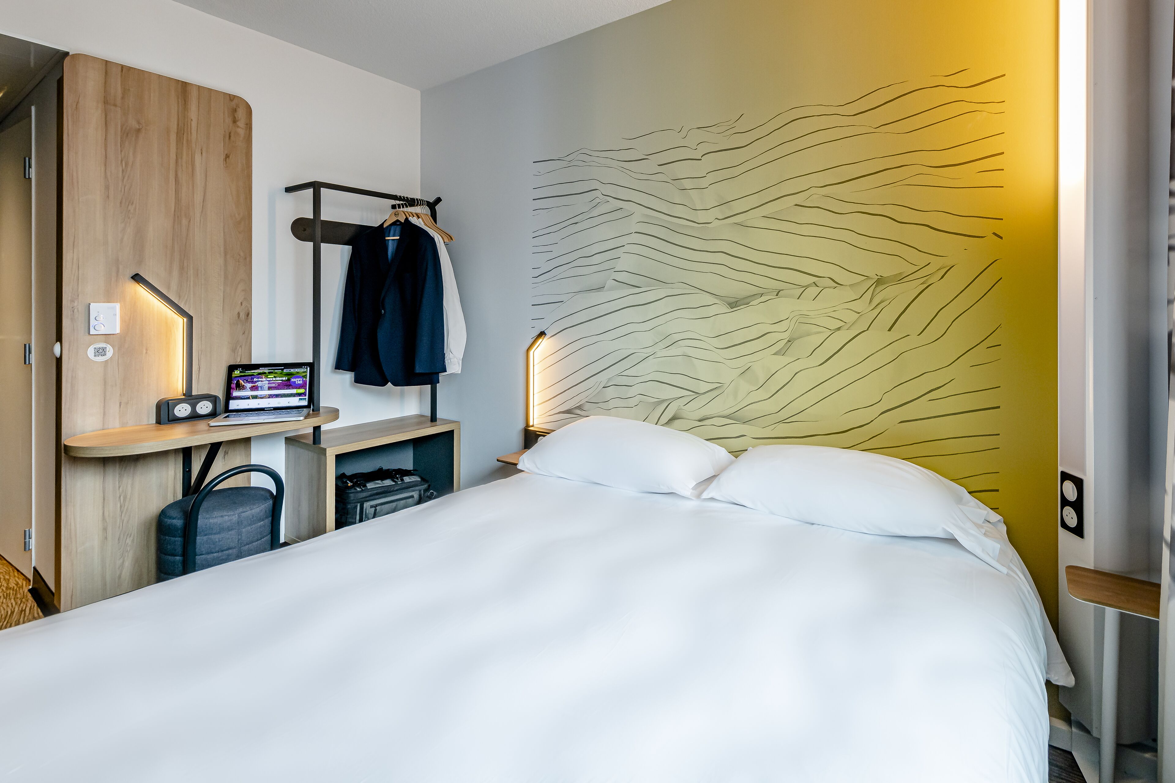 Habitació d'hotel moderna amb llit, escriptori amb ordinador portàtil, perguam i roba, i un disseny de paret únic.