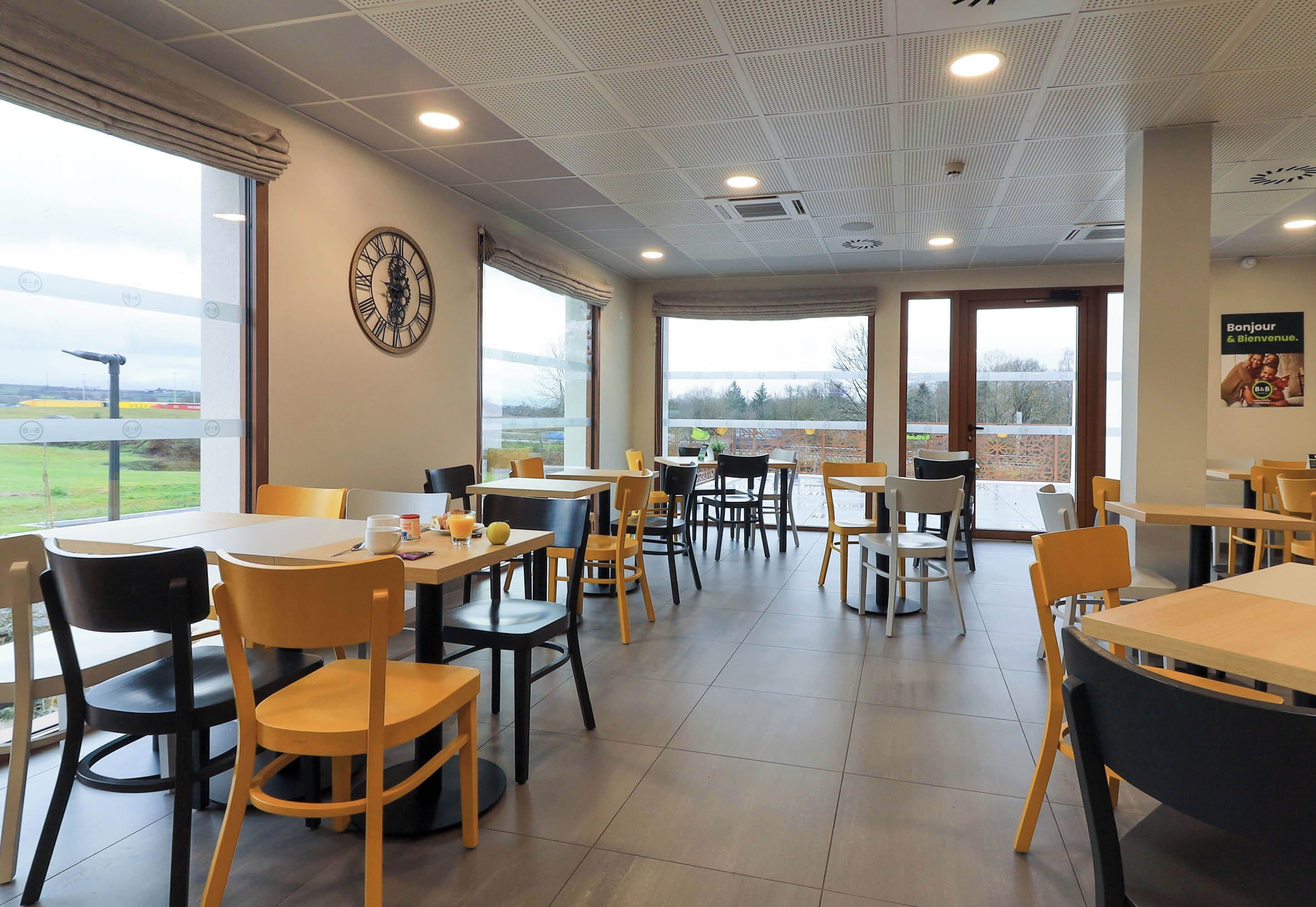 A spacious indoor area with multiple tables and chairs, large windows offering a view of the outdoors, and a clock on the wall.