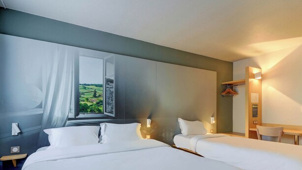Quarto de hotel com duas camas, uma janela ampla com vista para áreas verdes, uma mesa com cadeira e uma luminária fixada na parede.