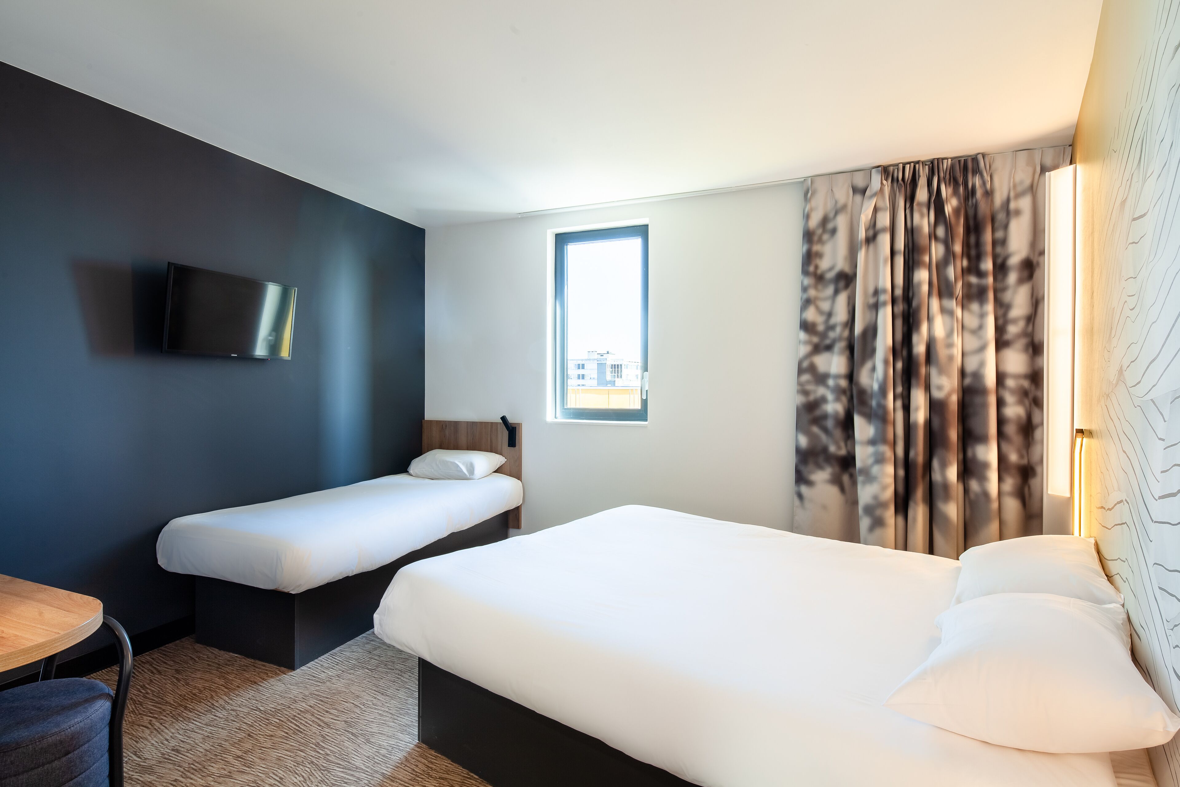 Habitación de hotel con una cama grande, una cama más pequeña, un escritorio, un televisor, una ventana y cortinas.