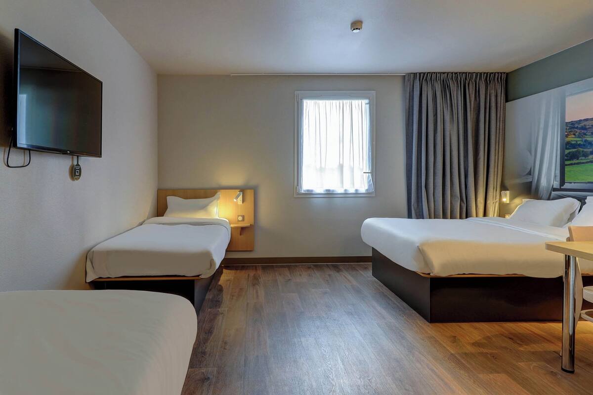 Ein Hotelzimmer mit zwei Einzelbetten, einem an der Wand befestigten Fernseher, einem Fenster mit Vorhängen und einem Holzboden.