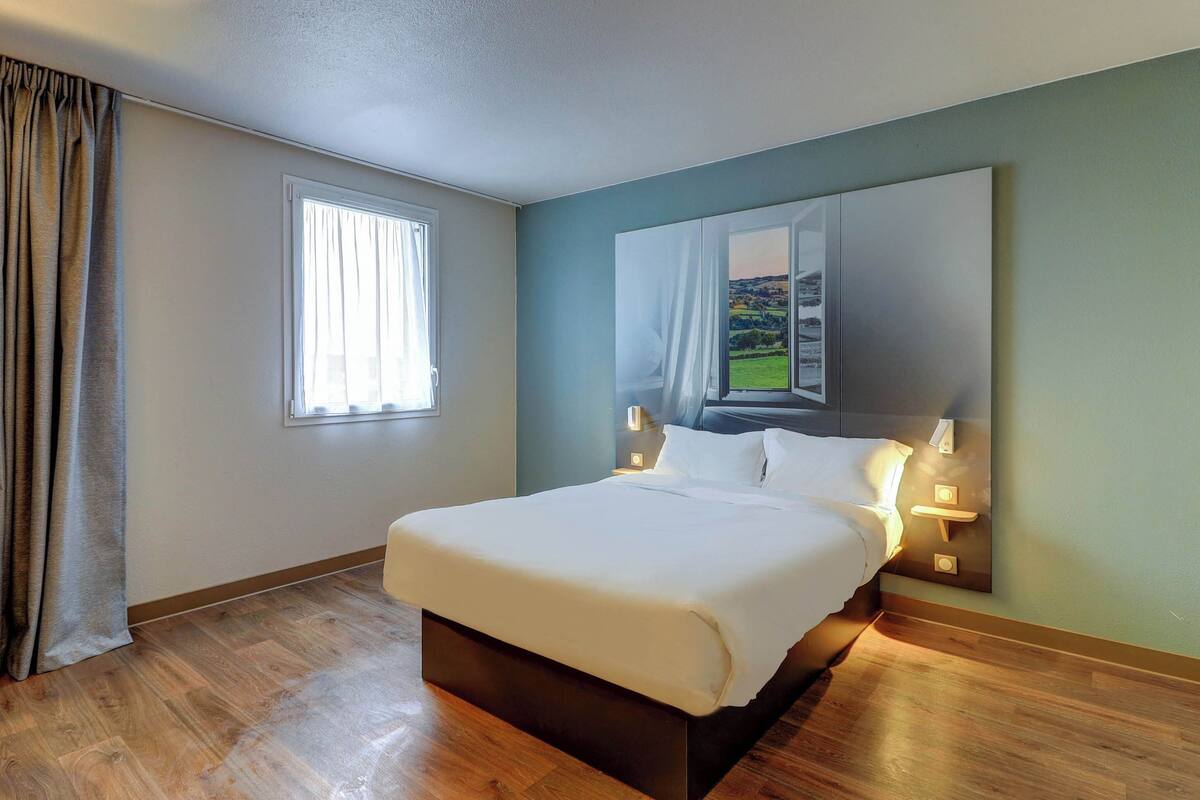 Ein Hotelzimmer mit einem großen Bett, einem Fenster mit Vorhängen, einem Nachttisch mit Lampe und einem Wandbild.