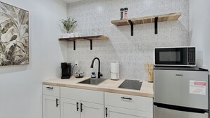 Una cocina compacta con gabinetes blancos, un refrigerador de acero inoxidable, un microondas, un lavamanos y una pequeña encimera. Hay dos estantes de madera sobre la encimera y una planta decorativa a la izquierda.