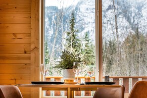 A wooden interior with a dining table set for two, a potted plant, and a mountain view through large windows.