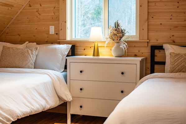 A cozy bedroom with two beds, a bedside table with a lamp, and a window with a view of trees.