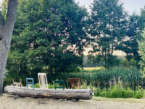Un espace extérieur rustique avec des sièges, comprenant un banc en tronc d’arbre, une chaise blanche, une chaise verte et une chaise rouge.