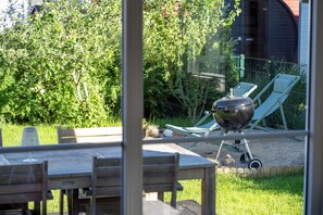 Een terras met een tafel, stoelen, een barbecue en uitzicht op een tuin met bomen en een hek.
