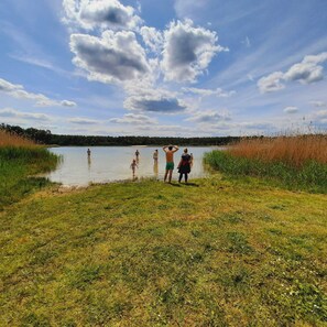 Un groupe de personnes se tient au bord d’un lac, avec une prairie au premier plan et une forêt en arrière-plan, sous un ciel partiellement nuageux.