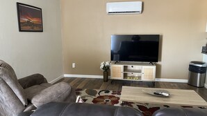 A living room with a beige wall, a mounted air conditioner, a flat-screen TV, a wooden coffee table, and a patterned rug. - 2 Bedroom Suite in Ridgecrest  (Ridgecrest)