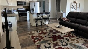 A modern kitchen with white cabinets, a microwave, and a refrigerator. There is a dining area with a table and chairs, a black leather sofa, and a patterned rug. - 2 Bedroom Suite in Ridgecrest  (Ridgecrest)