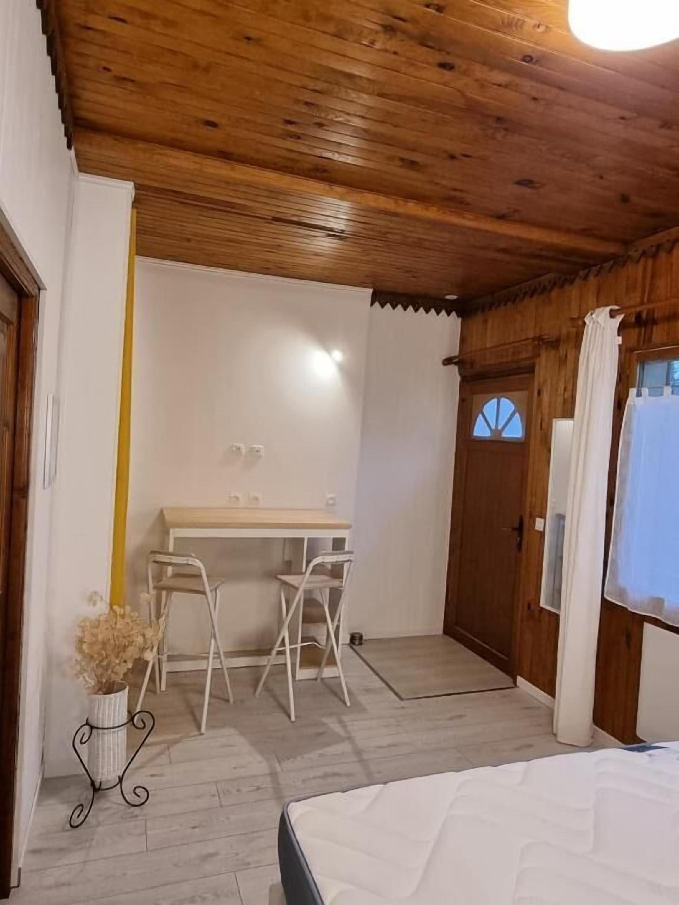 A bedroom with a wooden ceiling, a bed, a desk with two chairs, and a window with curtains.