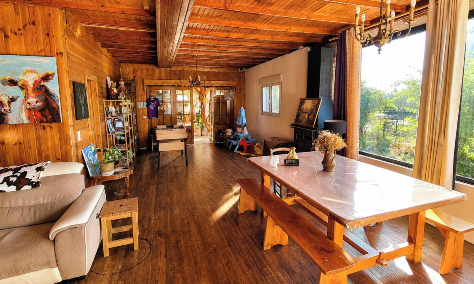 A cozy room with wooden walls and ceiling, a large wooden table with benches, a sofa, and a window with a view of greenery.