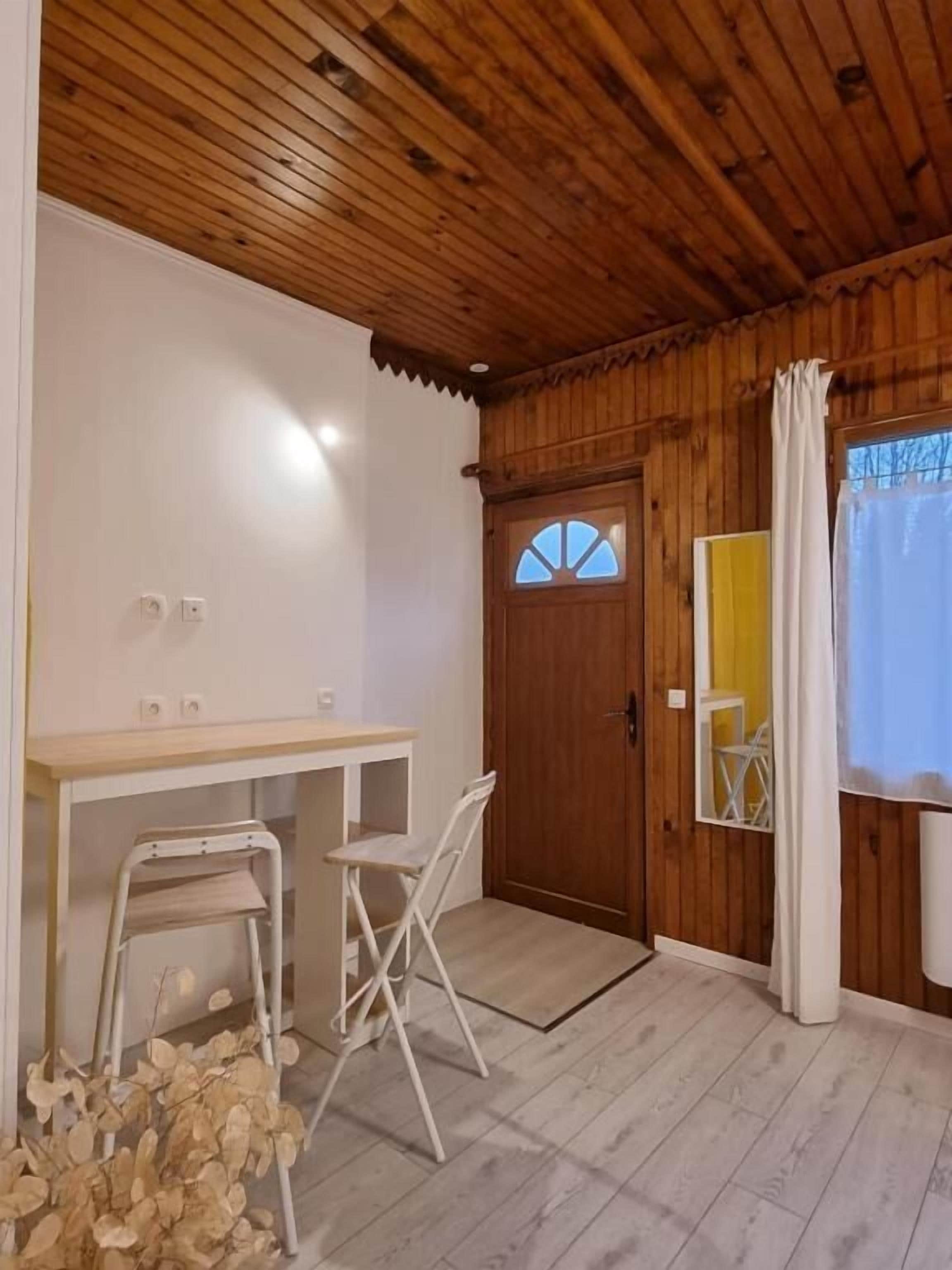 A room with a wooden ceiling, a wooden door with a window, a small desk with a chair, and a mirror.