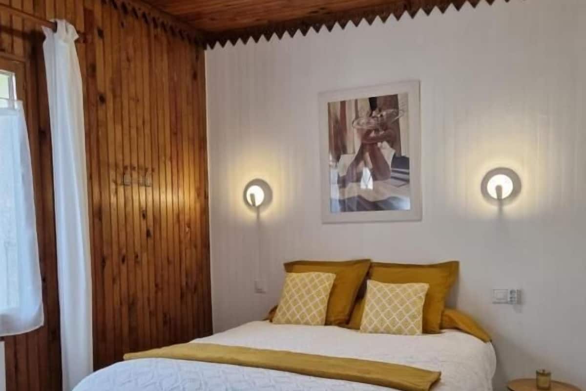 Une chambre avec un plafond en bois, un lit avec une literie blanche et jaune, deux lampes fixées au mur et un tableau encadré accroché au mur.