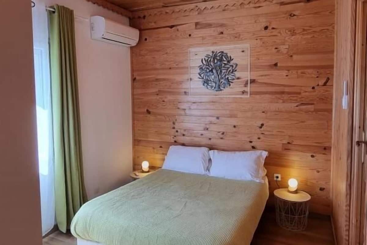 Une chambre à coucher aux murs lambrissés, avec un lit, deux lampes de chevet, un climatiseur fixé au mur et une fenêtre avec des rideaux.
