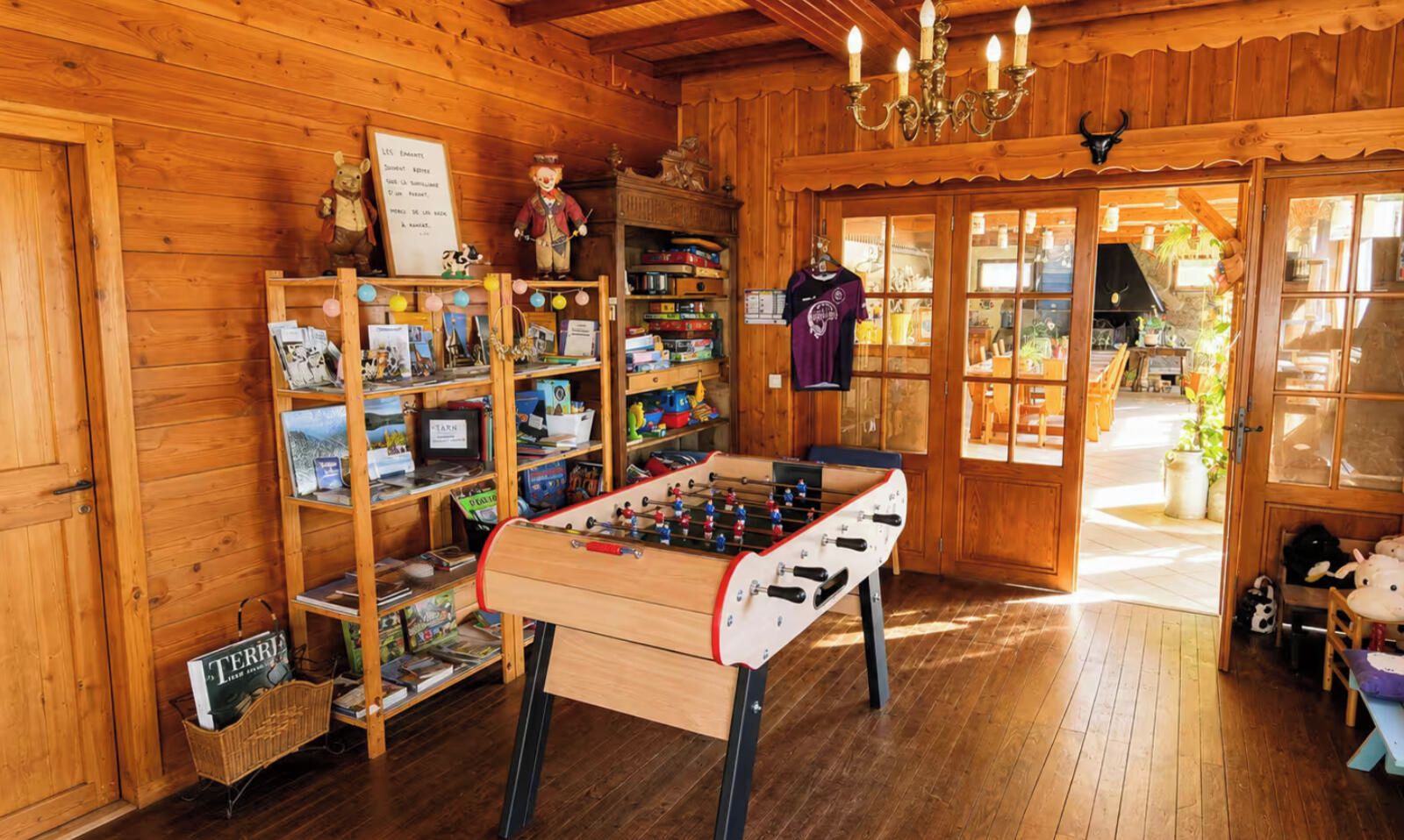 A cozy room with wooden walls, a foosball table, shelves with books and toys, and a door leading outside.