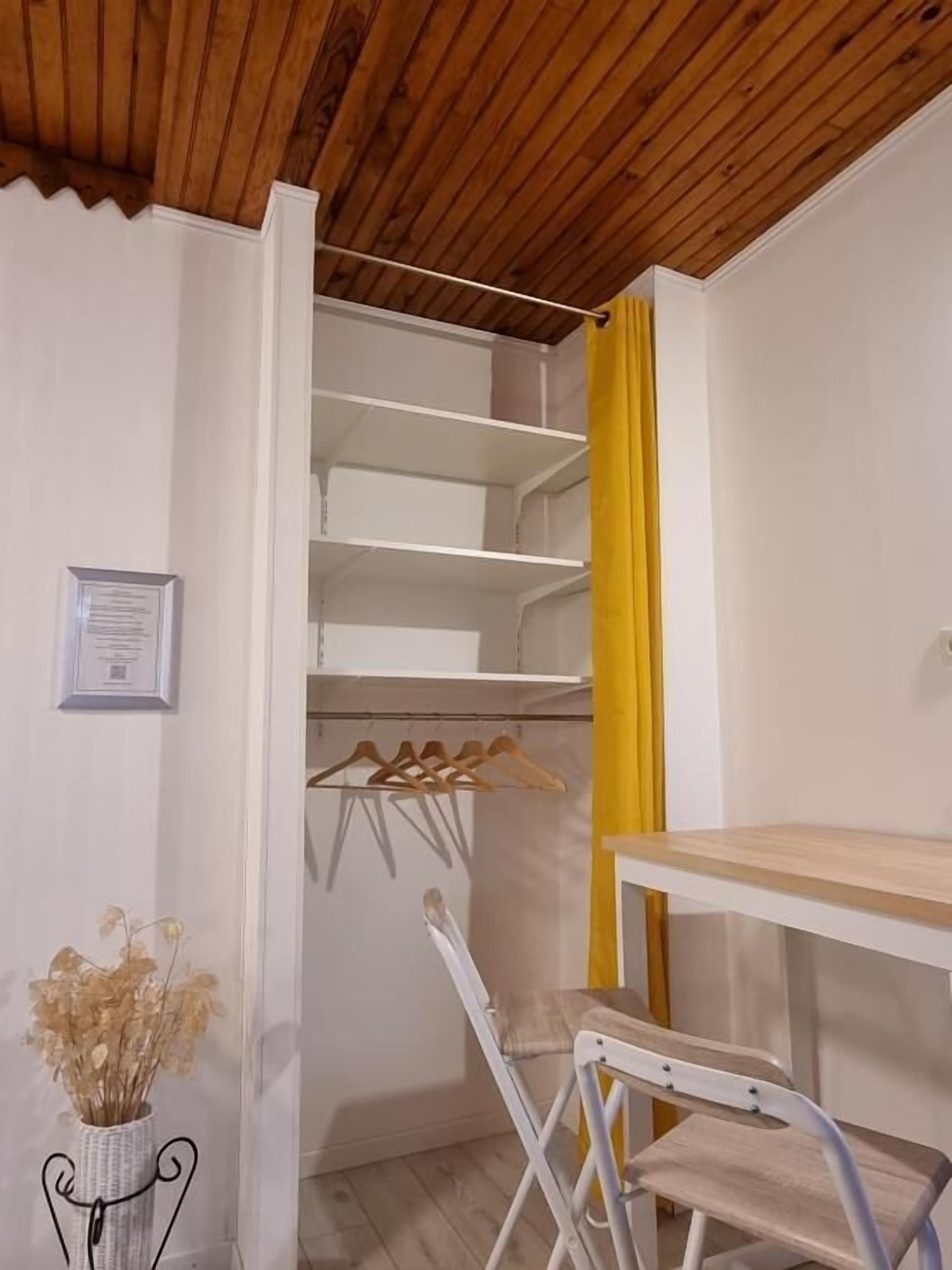 A room with a wooden ceiling, a white wall, a yellow curtain, a white desk, a white chair, and a metal hanger rack.