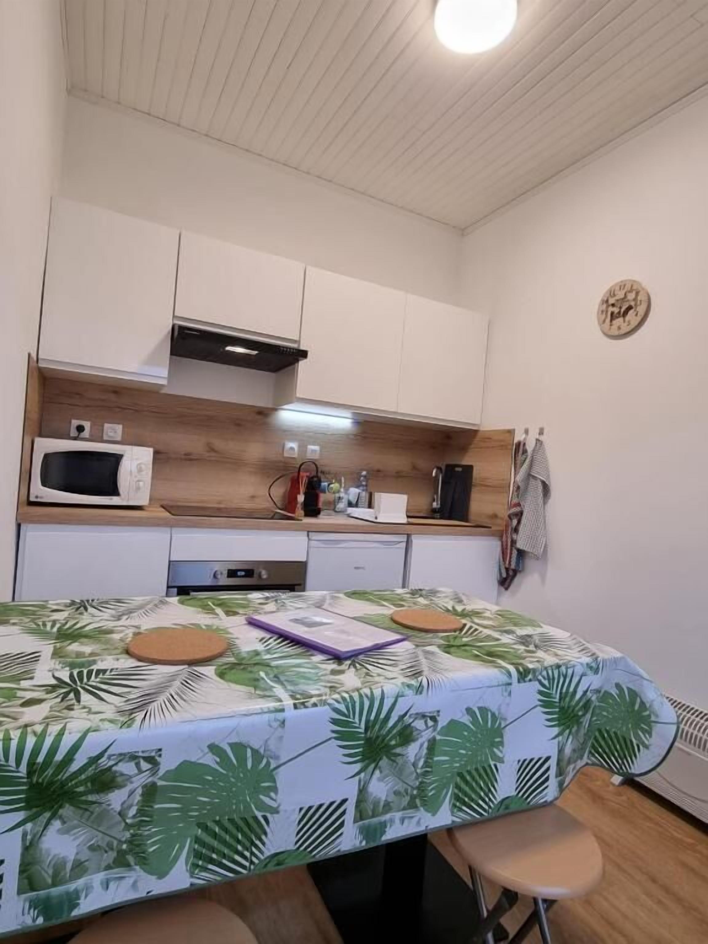 A compact kitchen with white cabinets, a microwave, and a small countertop. A table with a tropical leaf tablecloth and a notebook is in the foreground.
