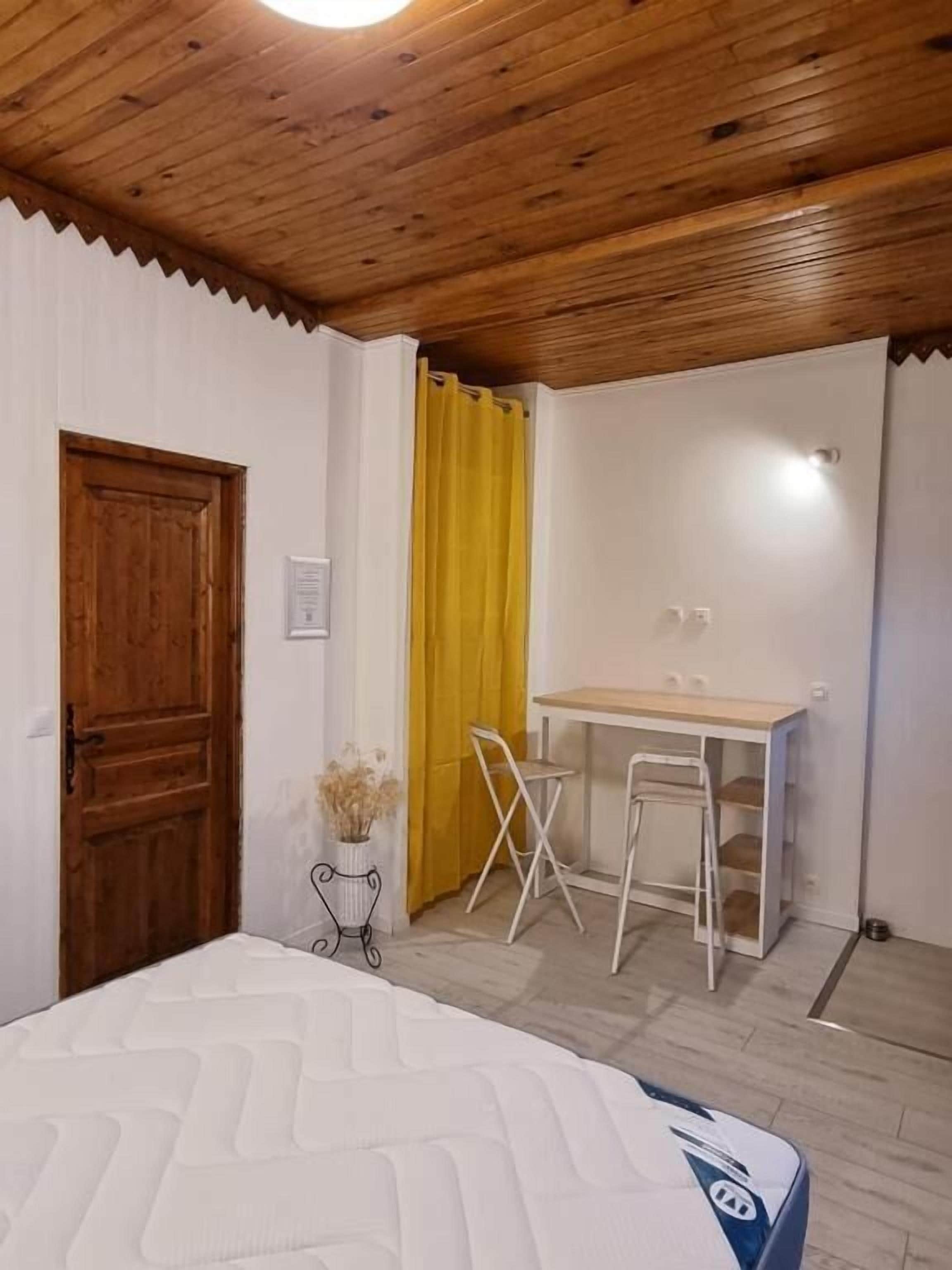 A bedroom with a bed, a wooden ceiling, a desk with a chair, and a yellow curtain.