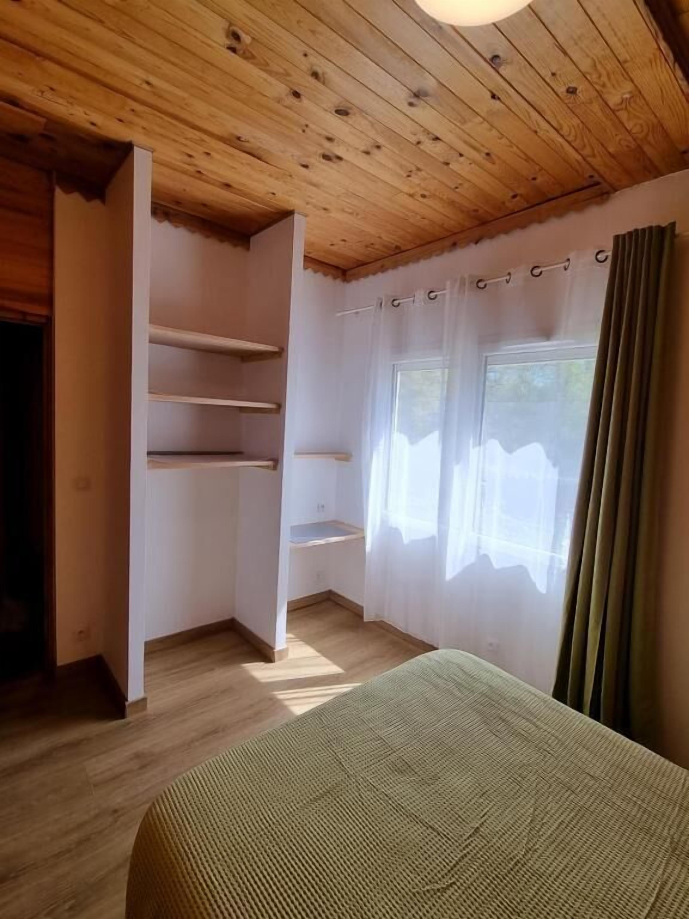 A bedroom with wooden ceiling, a bed, a window with curtains, and built-in shelves.