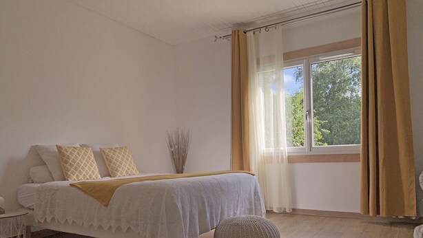 A bedroom with a bed, a bedside table, a window with curtains, and a view of greenery outside. - Les Tables Aux Vaches (La Salvetat-sur-Agout)