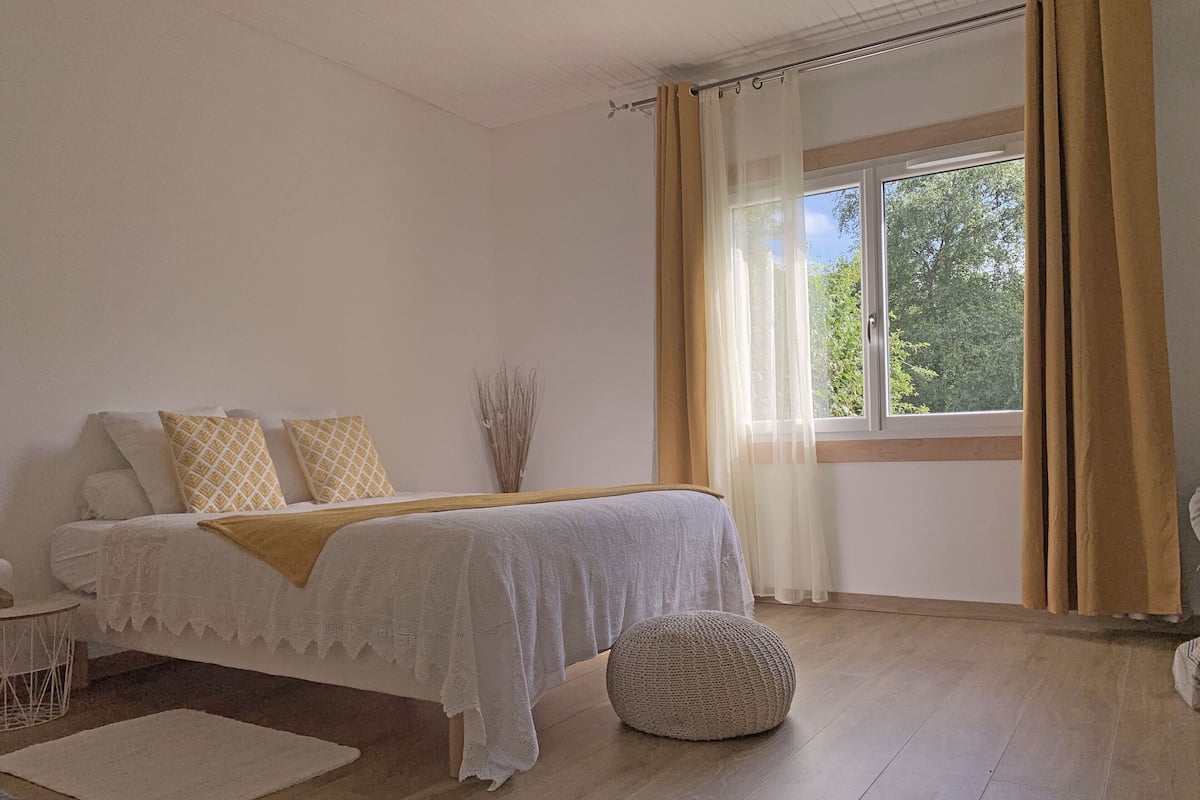 Une chambre à coucher avec un lit, une table de chevet, une fenêtre avec des rideaux et une vue sur la verdure à l’extérieur.